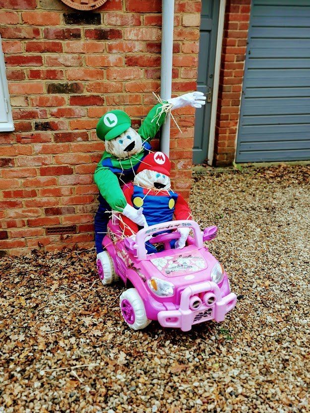 Scarecrow figures of Mario and Luigi in a pink toy car; Luigi is waving, set against brick wall.