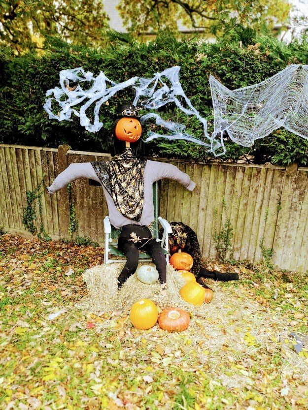 Scarecrow decoration with pumpkin head, seated in a chair, surrounded by pumpkins and cobwebs.