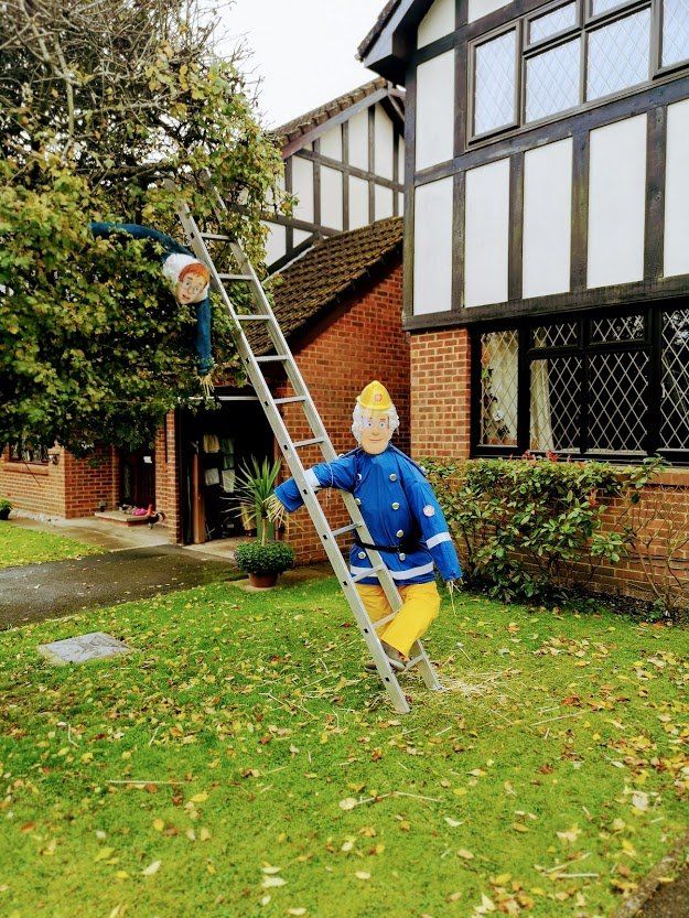 Two scarecrows dressed as firefighters on a ladder in front of a house.