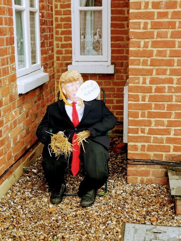 Scarecrow dressed in a suit, red tie, and straw hair, seated next to a brick wall.