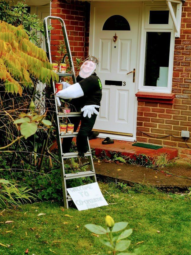 Scarecrow on a ladder near a house, holding cans. A sign sits on the lawn.