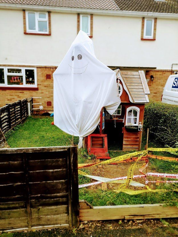 A ghost decoration made from a white sheet hangs in a front yard next to a playhouse, surrounded by caution tape.
