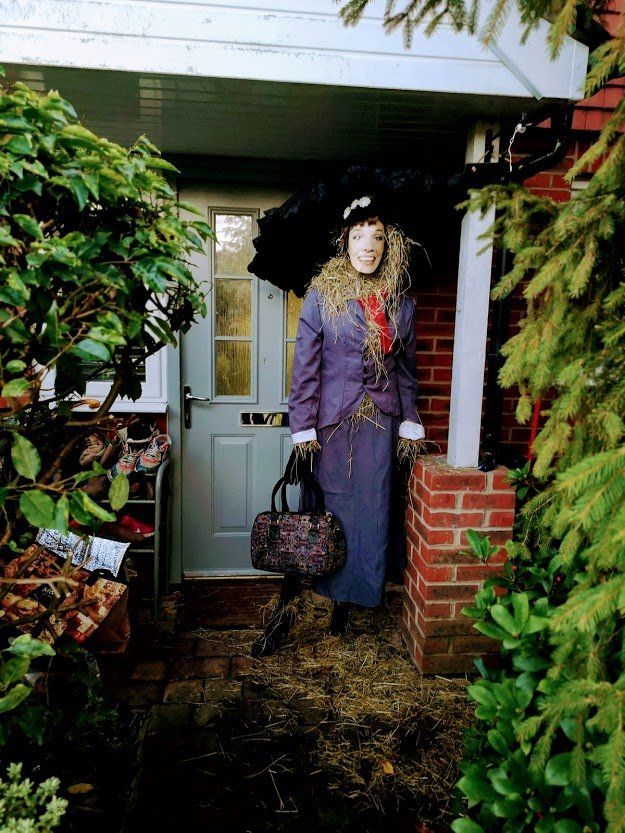 Person dressed as Mary Poppins stands in a doorway, holding a handbag and umbrella.