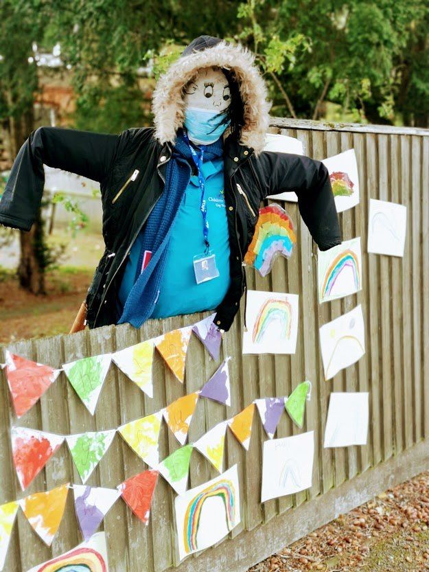 Scarecrow wearing a mask and jacket, adorned with rainbows and colorful bunting on a wooden fence.