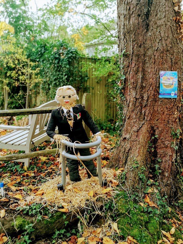 Scarecrow in a suit using a walker, near a tree, autumn leaves, and a bench.
