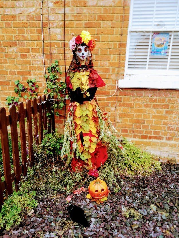 Scarecrow dressed for Day of the Dead, with floral crown and painted face, near a brick building. Pumpkin and bird at its feet.