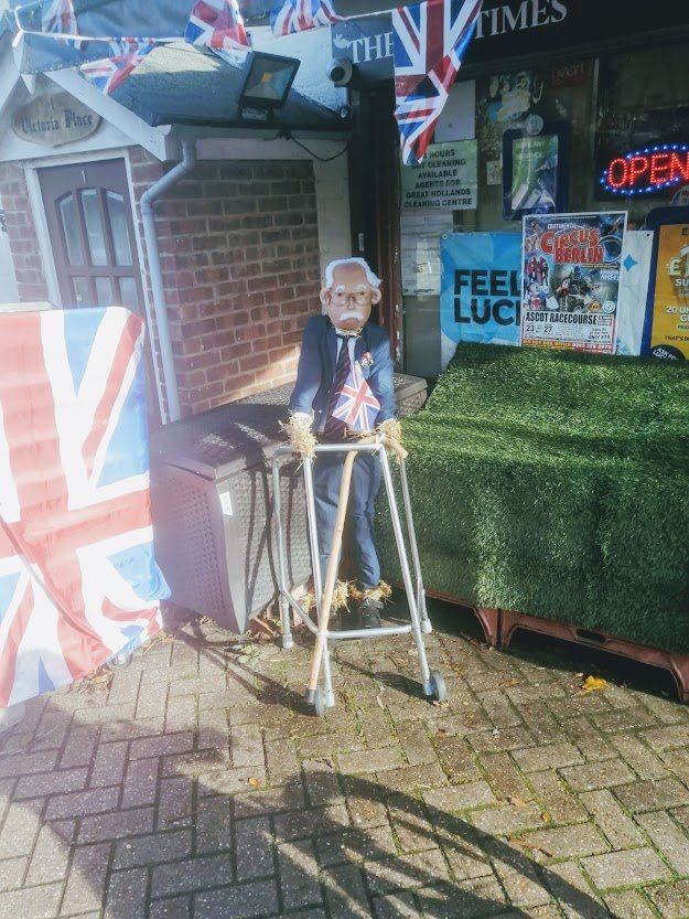 Statue of Captain Tom Moore with a Union Jack flag on a walker outside a building with flags.