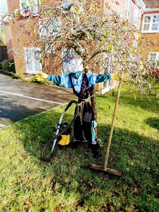 A scarecrow wearing clothes stands near a tree, lawnmower and broom on grass.