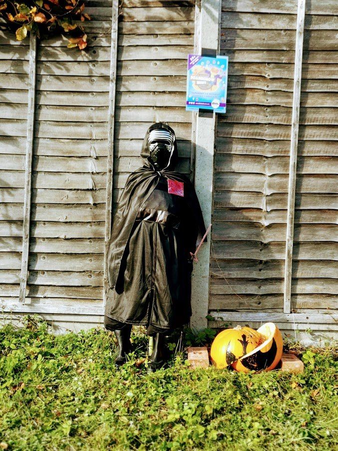 Scarecrow dressed in black, with a mask, in front of a wooden fence. A pumpkin sits at its feet.