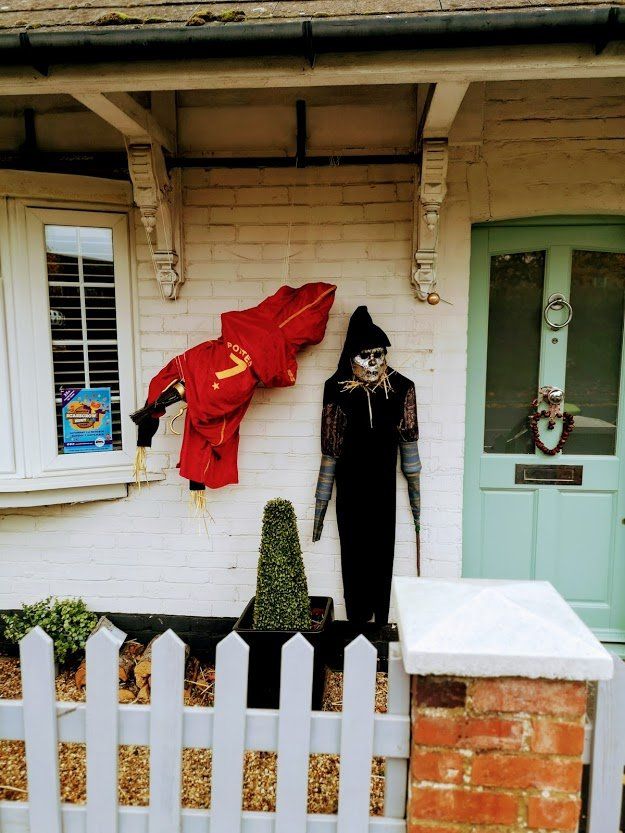 Two scarecrows stand outside a white brick house, one in red, one in black, next to a picket fence.