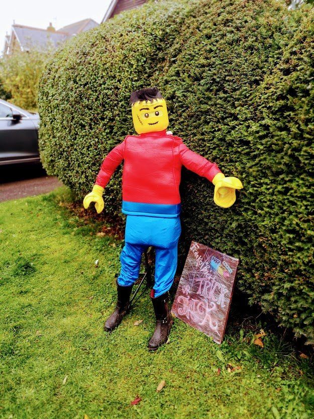 Lego-man scarecrow with yellow head, red shirt, blue pants, black boots, standing on grass near a hedge.