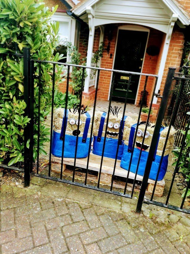 Three Minion-like figures made of blue plastic tubs, behind a black metal fence, in front of a house.