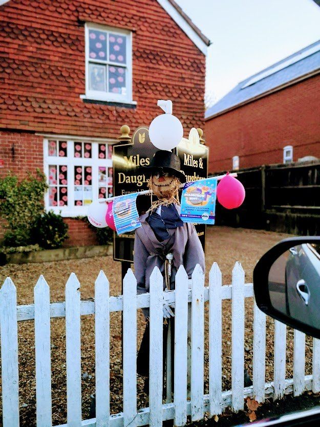 Scarecrow decorated with balloons in front of a building with a white picket fence.