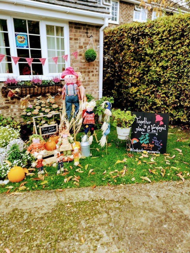 House front yard decorated for fall: scarecrows, pumpkins, colorful flowers, and a sign.