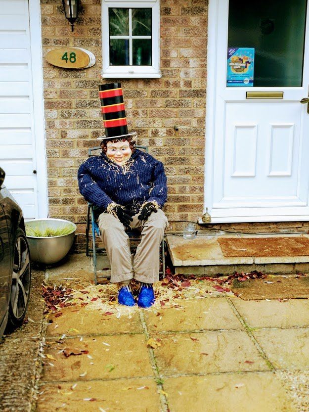 Scarecrow in a top hat seated on a chair in front of a house, number 48. Wearing blue shoes and dark blue jacket.