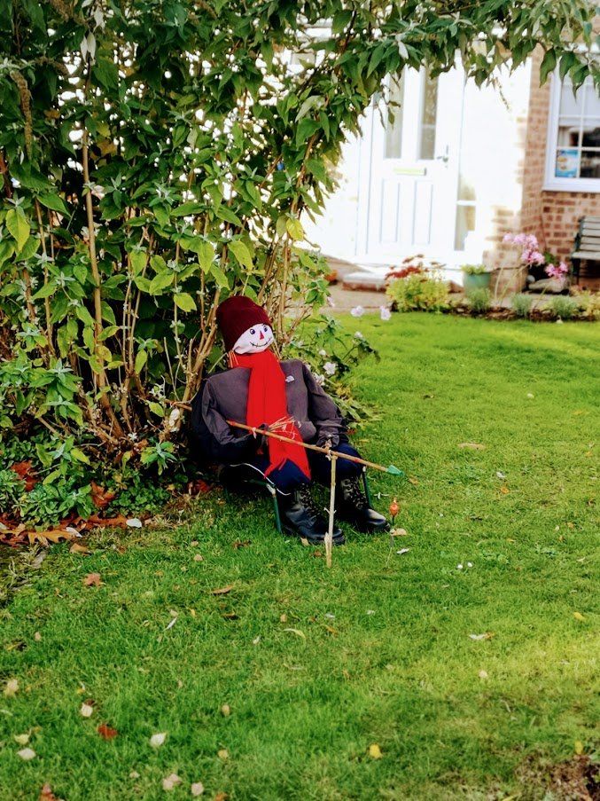 Scarecrow in a red hat and scarf, leaning against a bush in a garden.