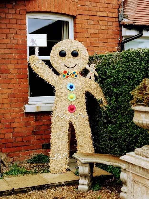 Large straw gingerbread man with button details stands by a brick building.