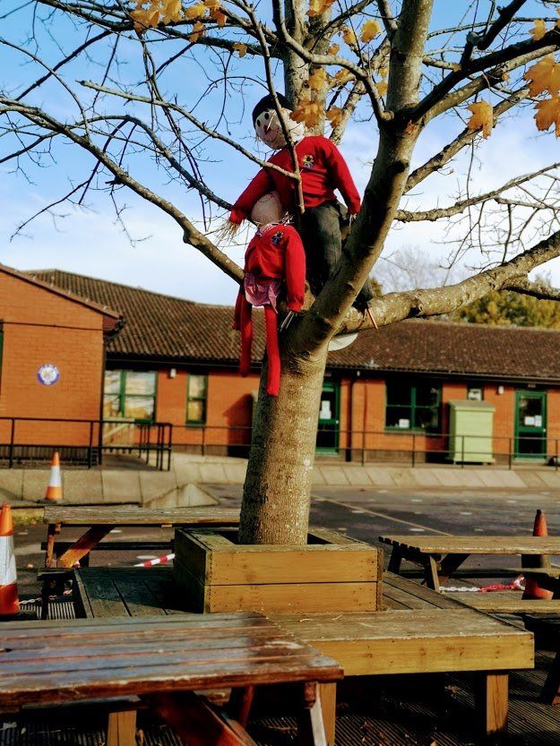Child in red climbs a tree in a schoolyard, near picnic tables and a brick building.