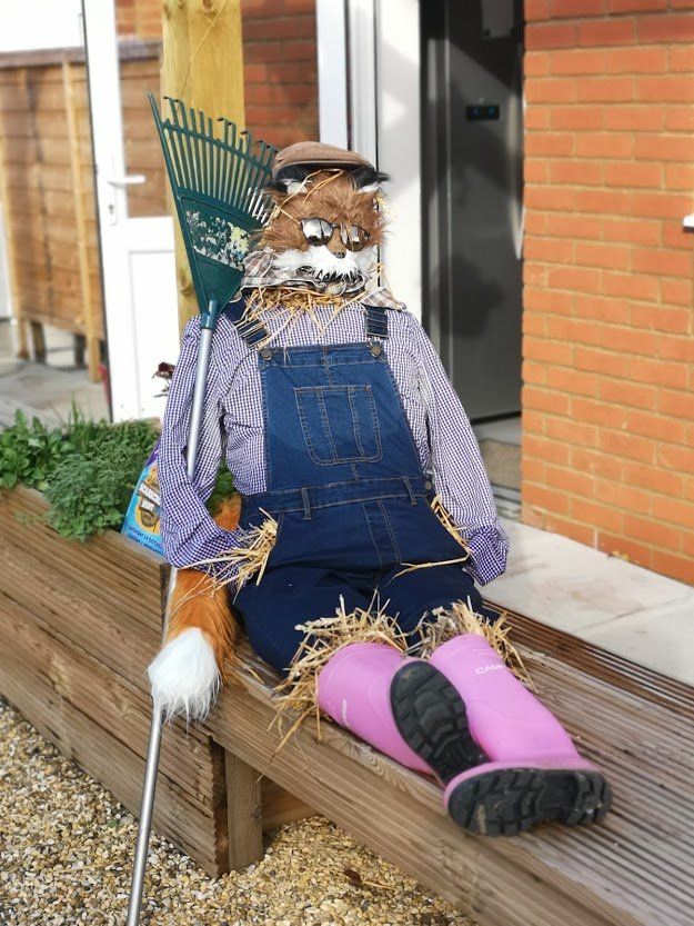 Fox scarecrow wearing overalls and boots, holding a rake, sitting on a wooden bench.