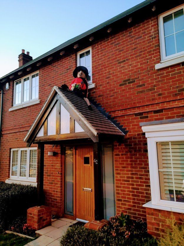 A figure wearing a hat sits on a porch roof of a brick house.