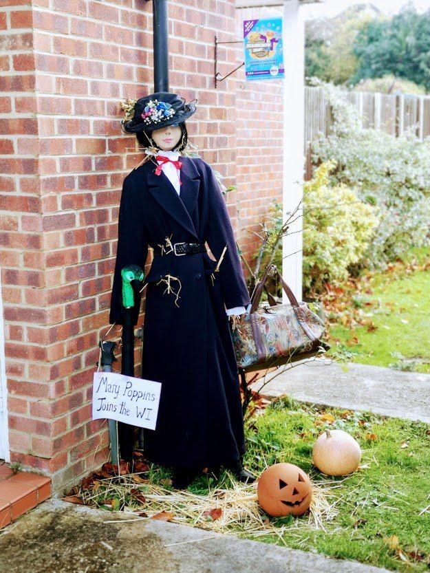 Scarecrow dressed as Mary Poppins with pumpkins in a yard.