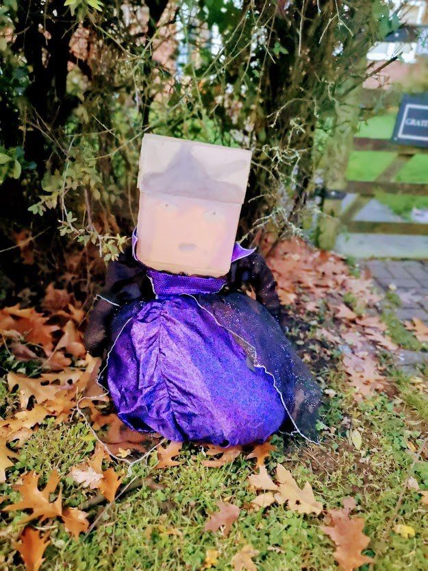 Halloween decoration of a figure with a paper bag head, purple skirt, and black netting, in a yard.