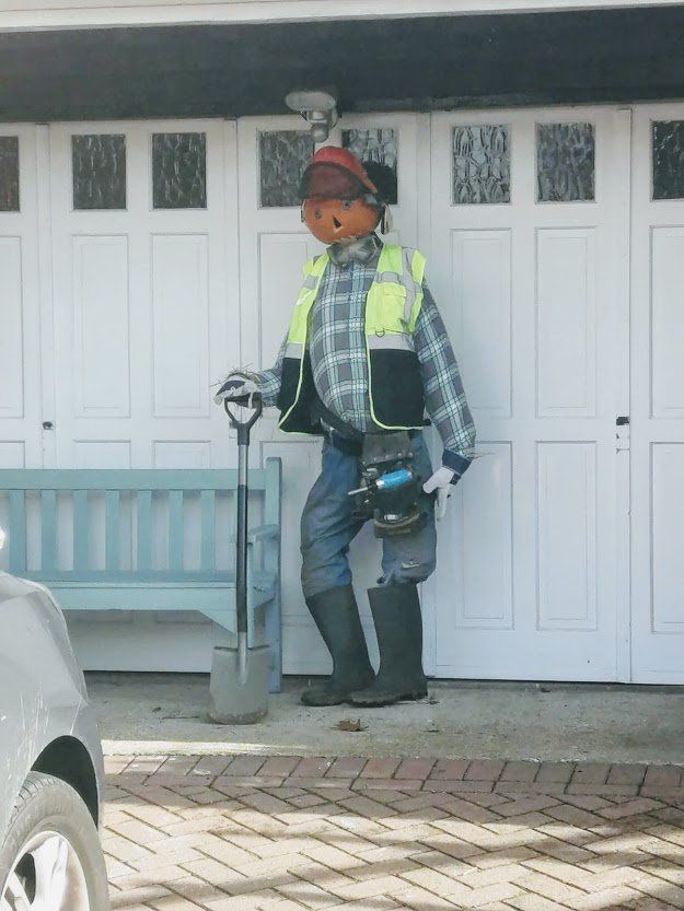 A scarecrow dressed as a construction worker stands in front of a white garage door, holding a shovel and tool.