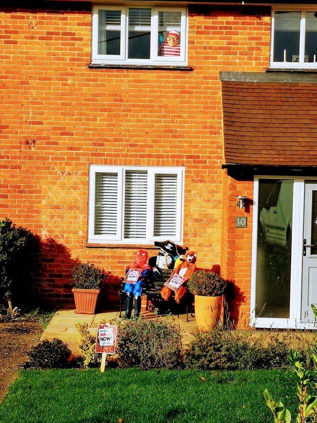 Red brick house with garden and two scarecrows on a bench. One window shows a person.