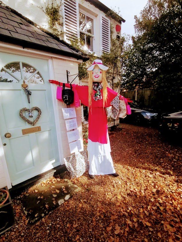 Scarecrow dressed in pink and white clothing stands in front of a house with a turquoise door.