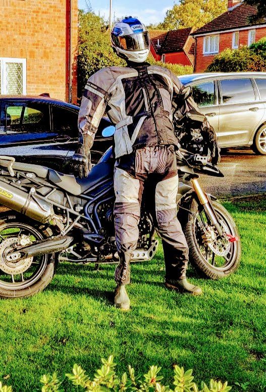 Person in motorcycle gear stands next to a Triumph motorcycle on grass. Houses and vehicles in the background.