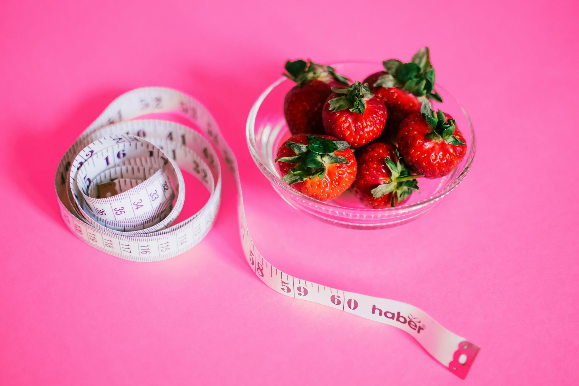 Pink background with glass bowl of strawberries and white measuring tape.