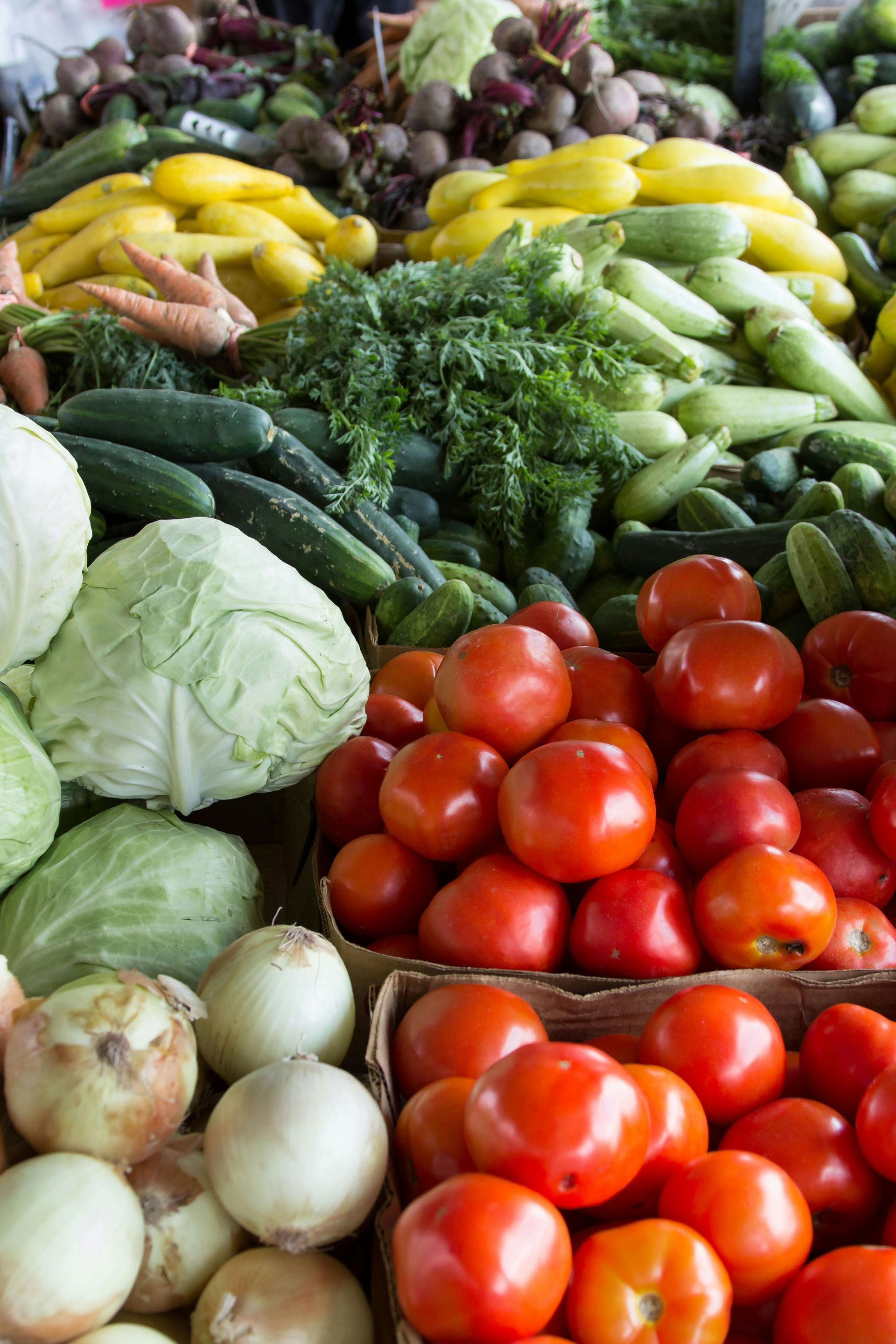Fresh produce at a farmer's market, including tomatoes, cucumbers, cabbage, onions, and squash.