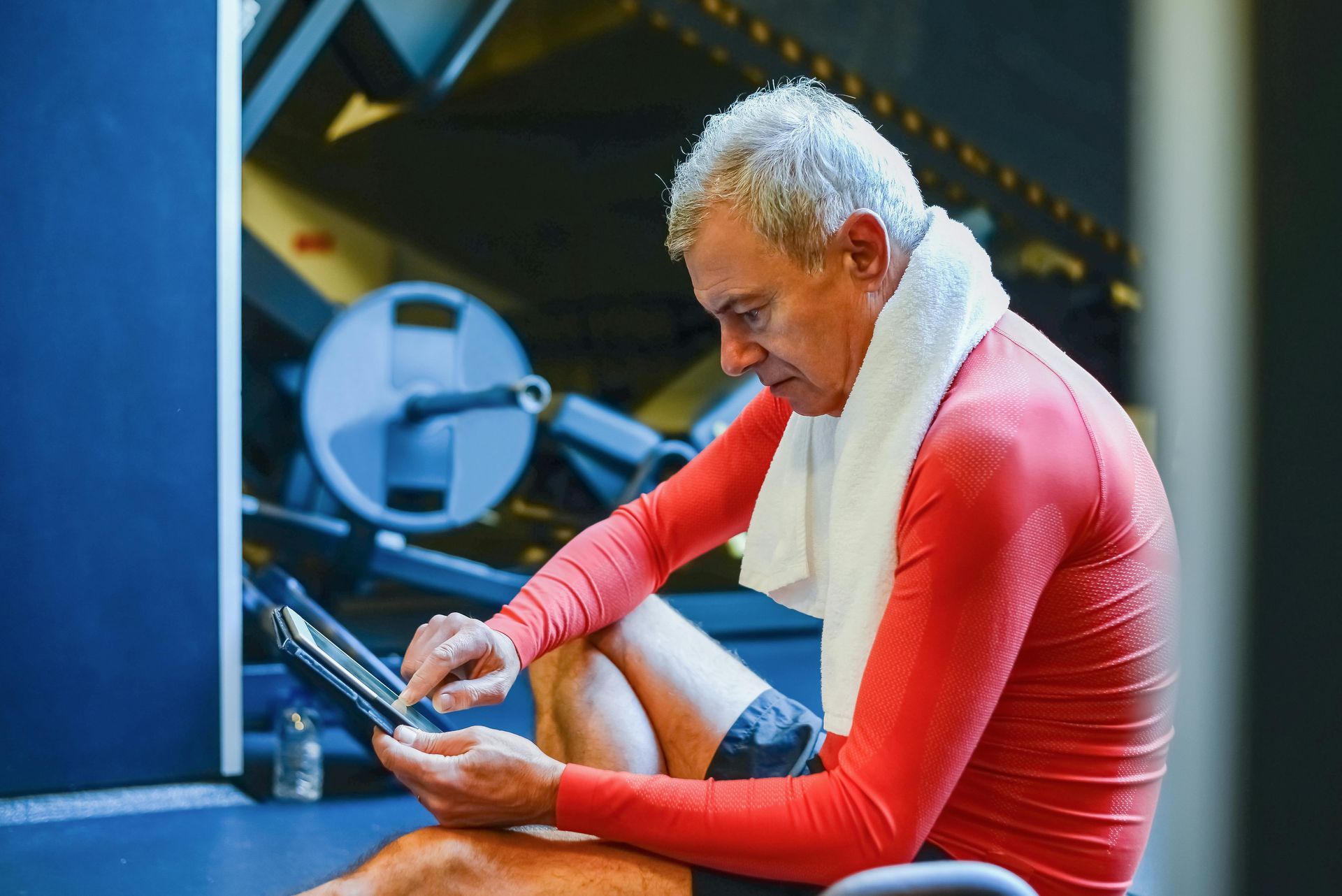 A man is sitting on the floor in a gym looking at a tablet.