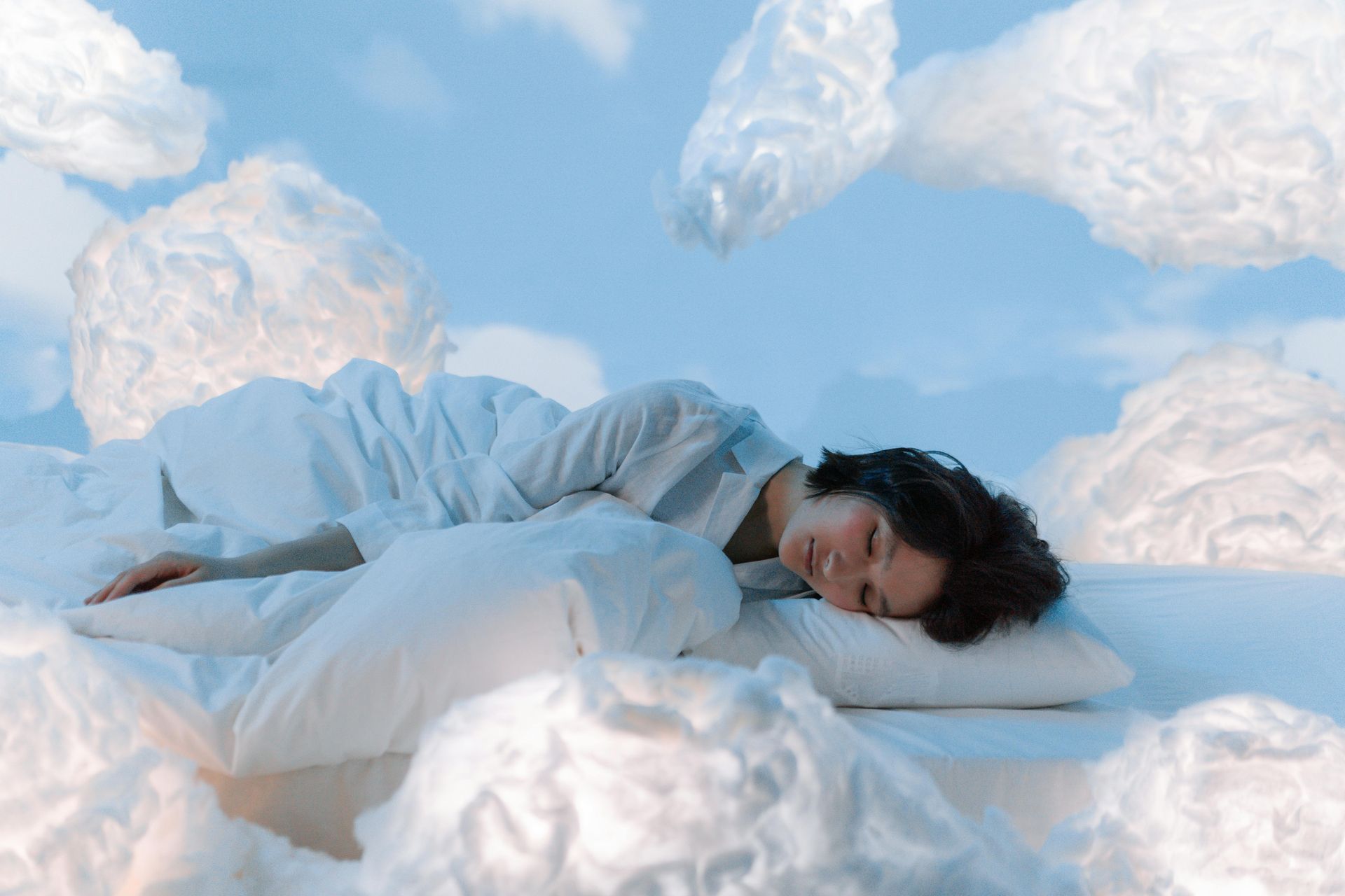 Woman sleeping peacefully in bed, surrounded by fluffy clouds in a sky-blue setting.