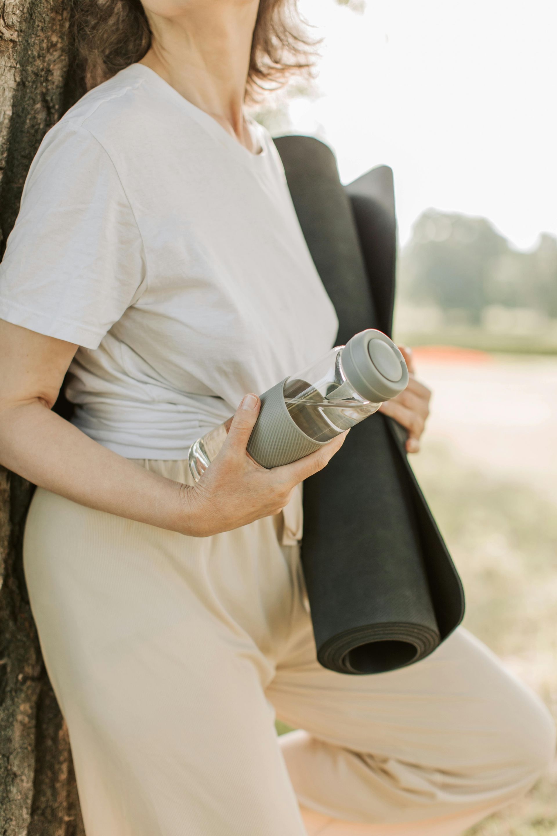 A woman is leaning against a tree holding a yoga mat and a water bottle.