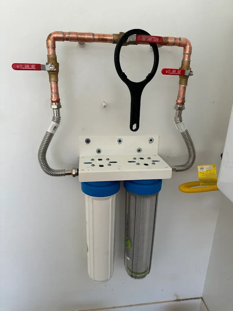 A wall-mounted dual water filtration system with copper piping, shut-off valves, and a black filter wrench hanging above.