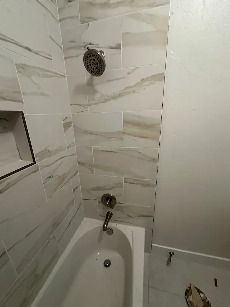 A bathroom shower and tub area featuring beige and white marbled wall tiles, a showerhead, faucet, and a recessed shelf.