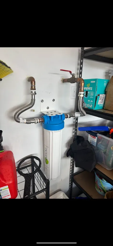 A white and blue water filtration system mounted on a garage wall with connected metal plumbing pipes and a shut-off valve.