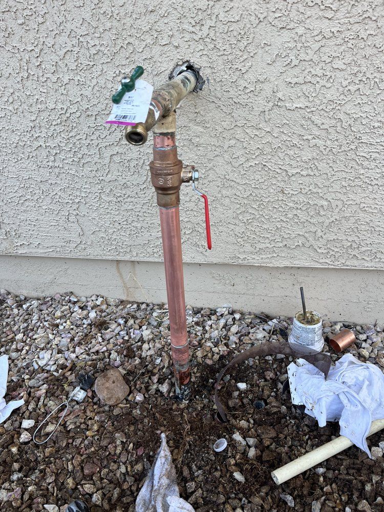 Outdoor copper plumbing fixture with a green handle and red shut-off valve installed against a textured stucco wall.