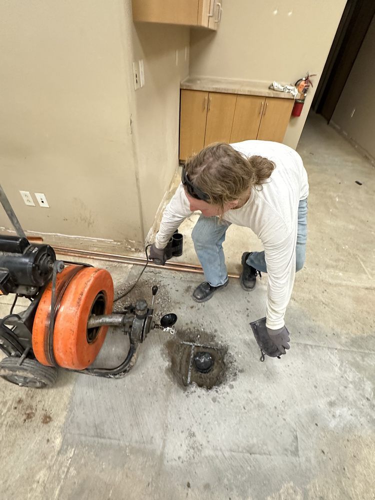 A technician in a white long-sleeve shirt uses a large orange industrial pipe cleaning machine on a concrete floor.
