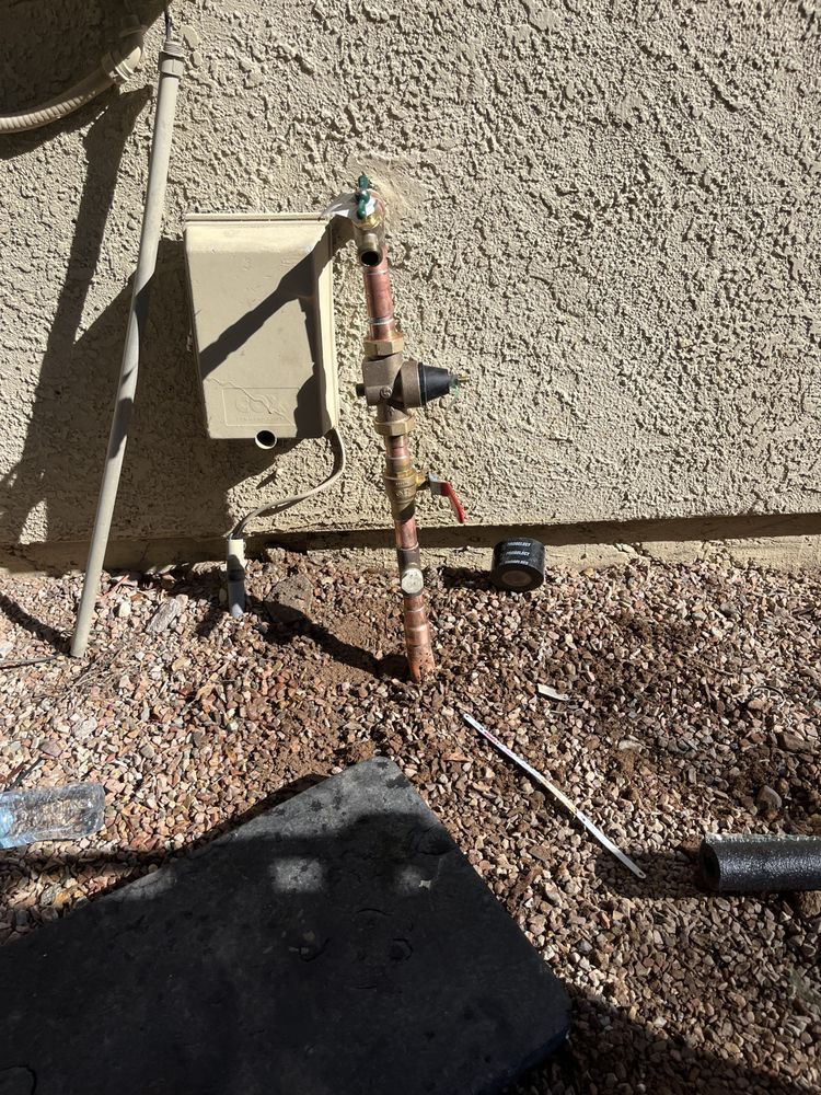 Copper plumbing pipes and a sprinkler control box mounted on an exterior stucco wall above dirt and landscaping rocks.