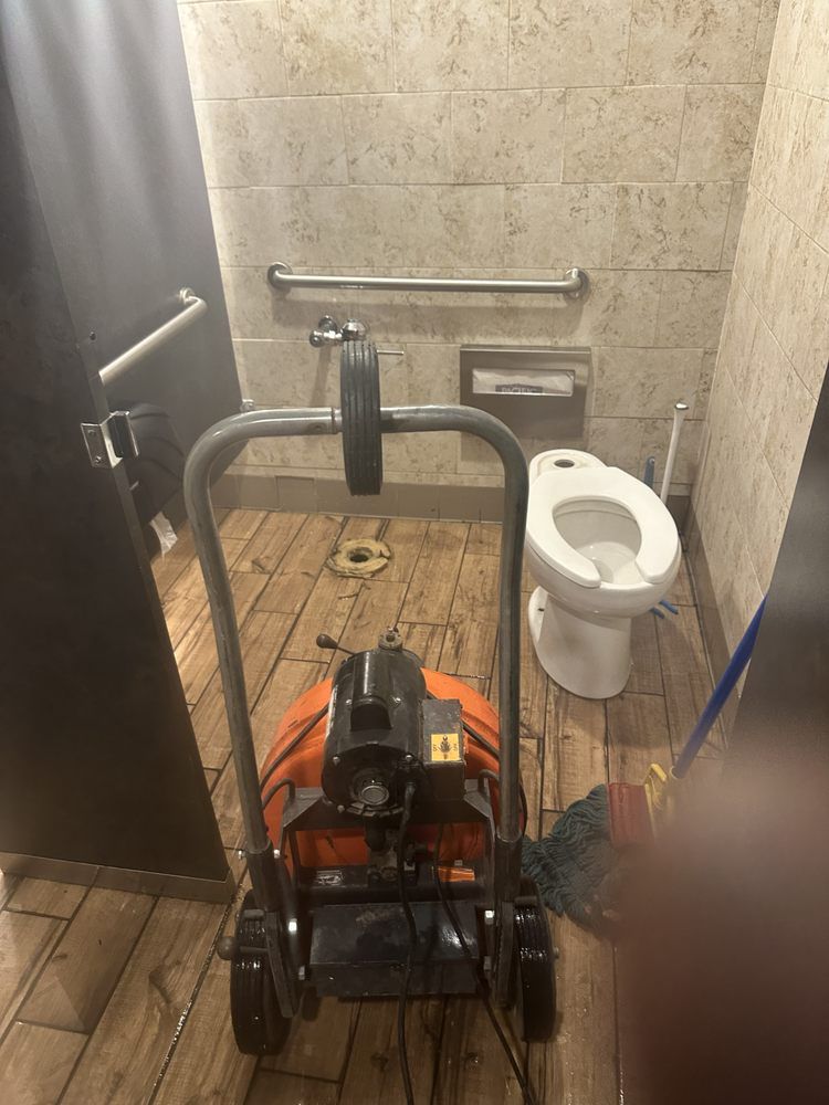 A toilet stall undergoing maintenance with a drain cleaning machine positioned on the floor in front of a detached toilet.