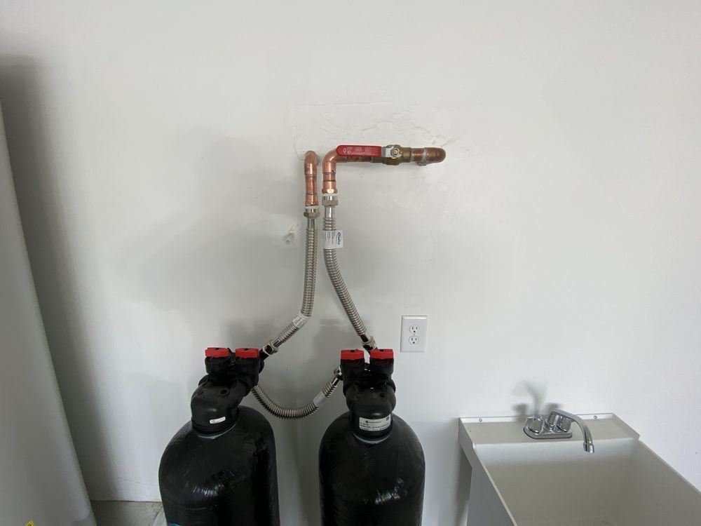 Two black water filtration tanks connected by braided metal hoses to copper pipes with a shut-off valve on a white wall.