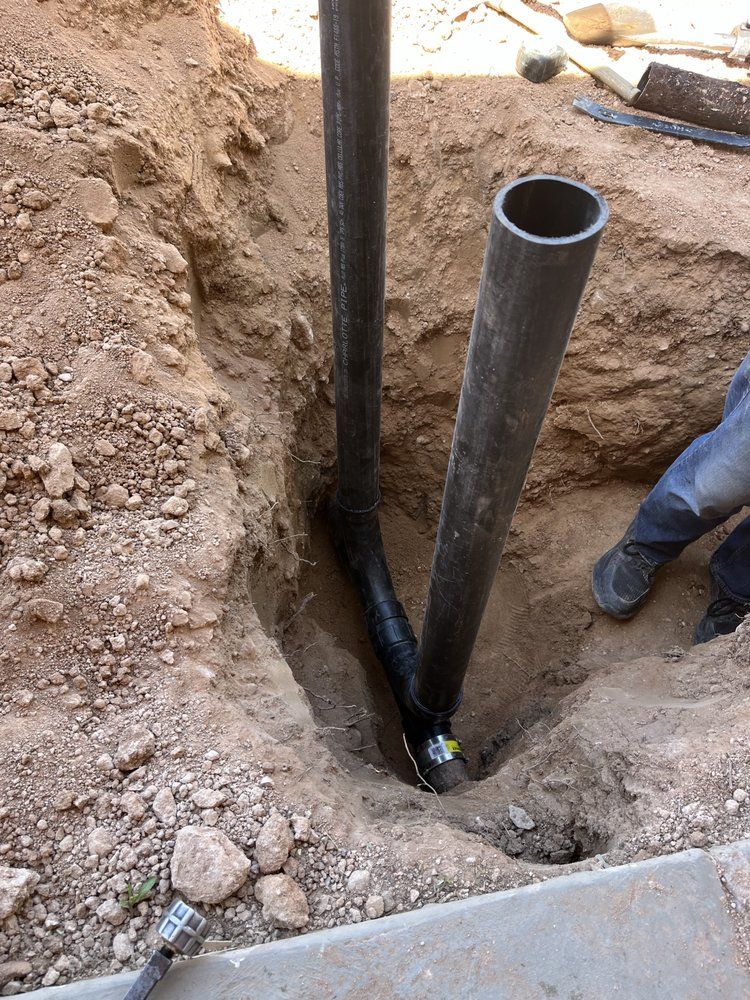 A deep trench in dirt containing two dark vertical pipes connected by an angled fitting, with a person’s feet visible.