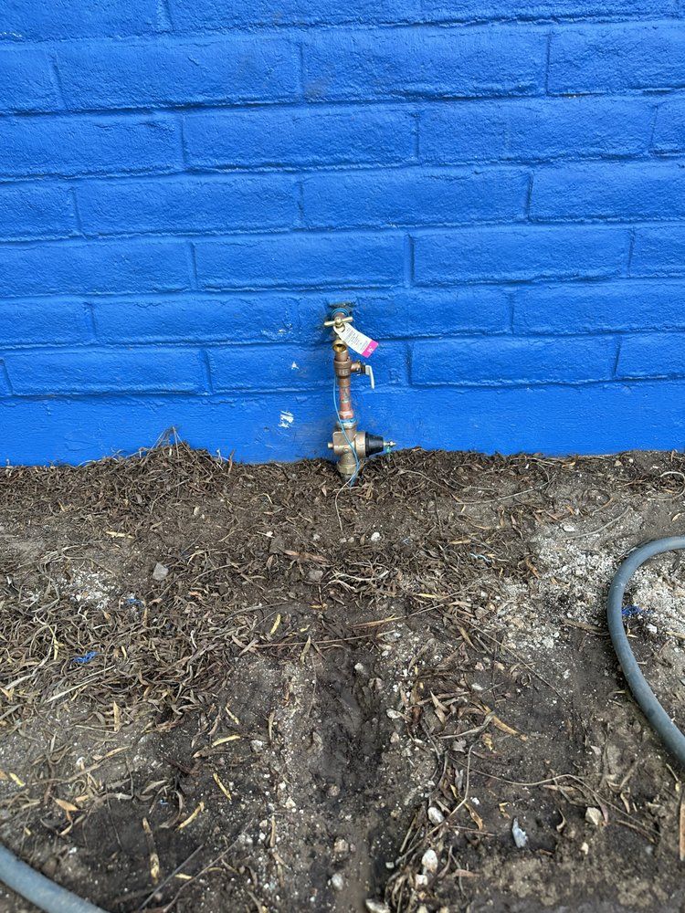 A vertical metal pipe with a faucet and a tag extends from a bright blue brick wall above a patch of dirt and mulch.