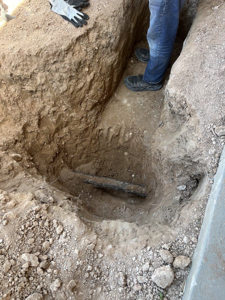A person in jeans and work boots stands beside a narrow trench in the ground containing an exposed pipe.
