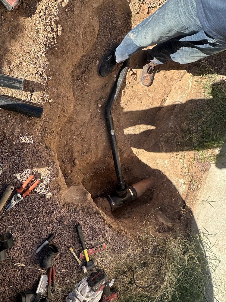 A person stands in a dirt trench while installing a new black pipe connection to a larger underground pipe.