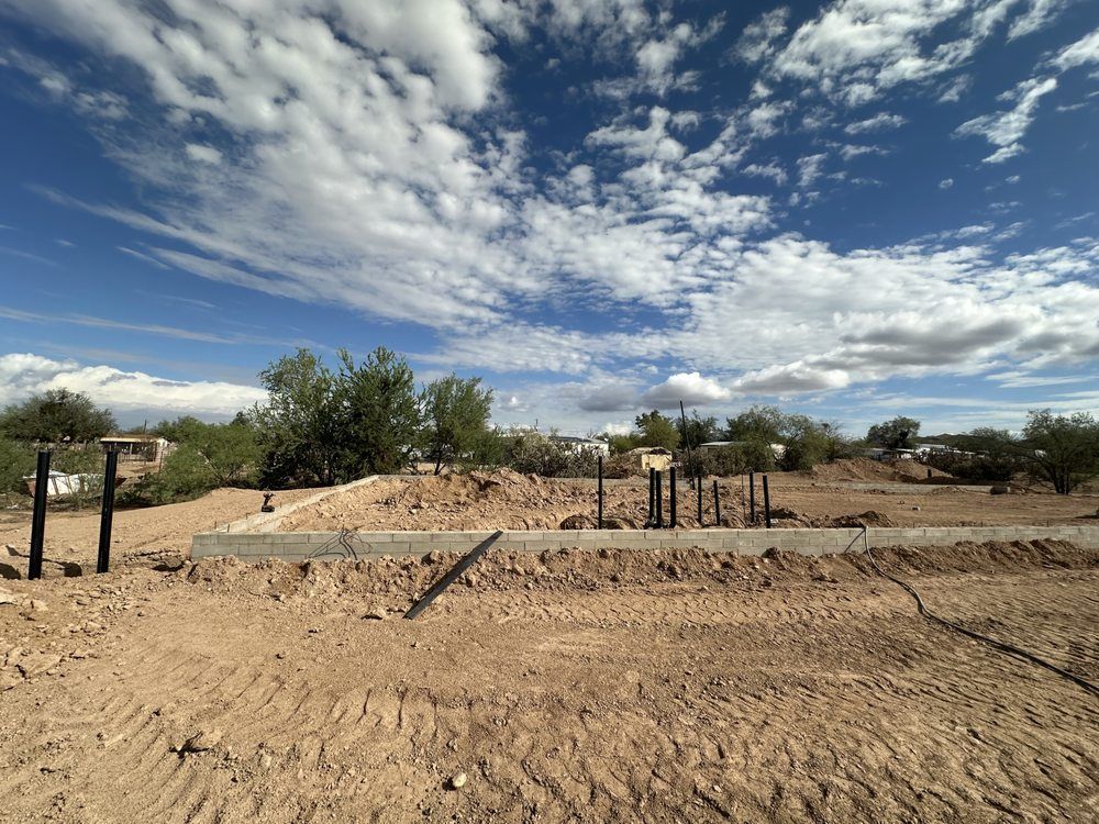 A construction site with a concrete foundation and metal posts set in a dirt lot under a cloudy, bright blue sky.