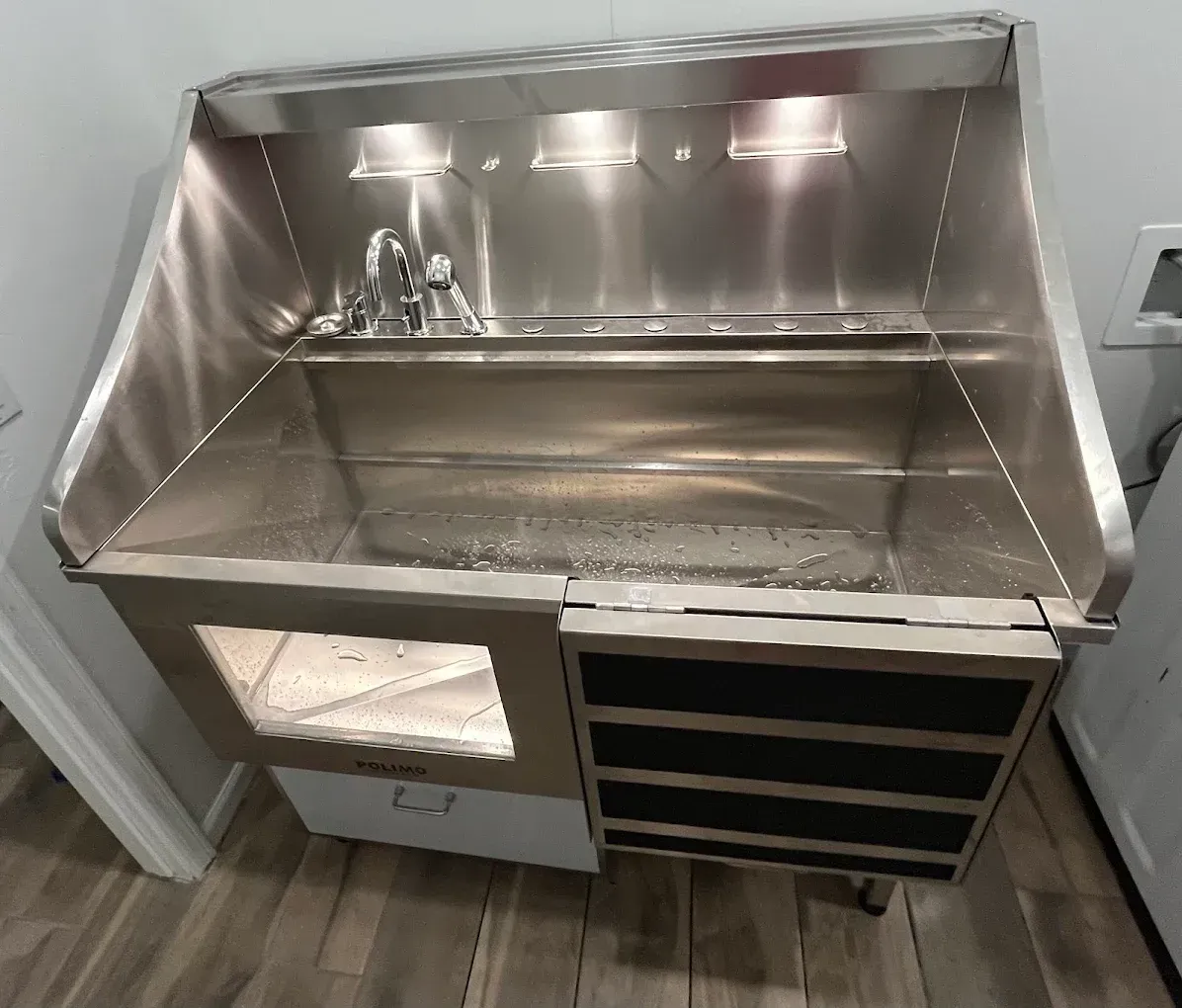 A stainless steel dog grooming tub with a side door and drawer unit, set against a white wall and wood-look floor.