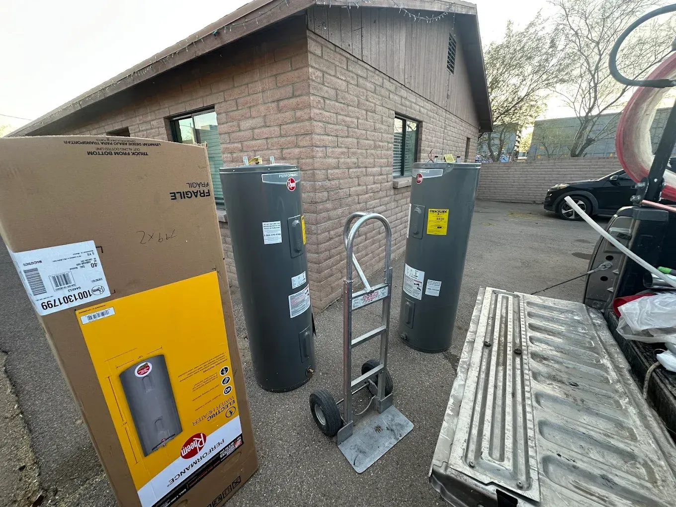Two grey water heaters and a hand truck sit on a driveway next to a cardboard water heater box and an open truck bed.